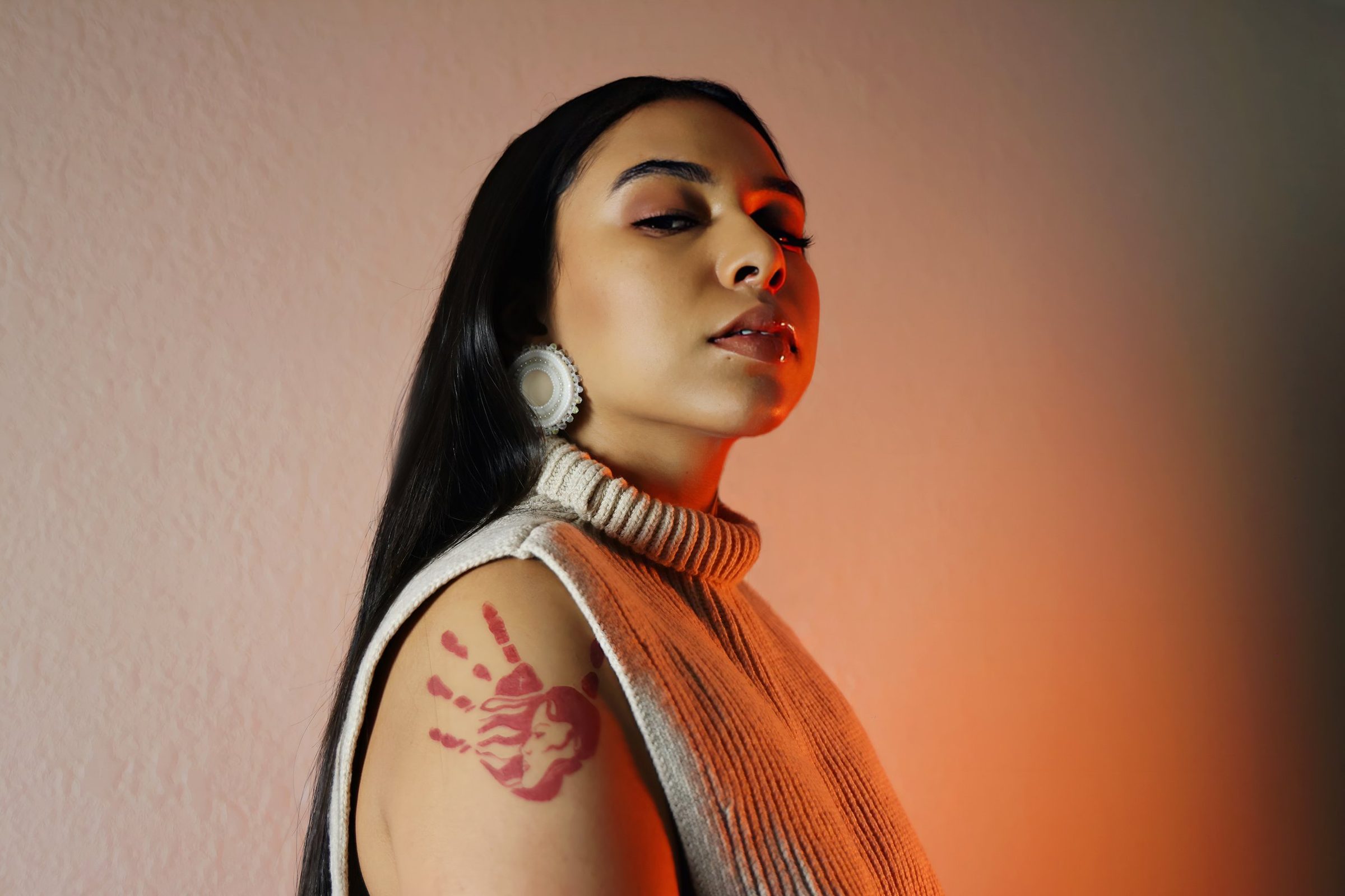 Stella Standingbear photographed against a warm orange-lit wall, the red MMIW handprint visible on her arm